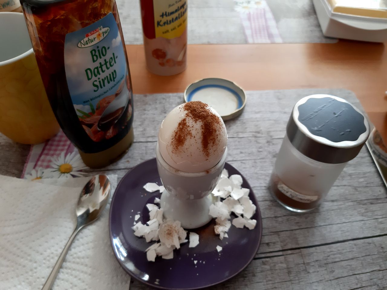 Ein gekochtes Frühstücksei im Eierbecher, dick mit Zimt bestäubt, daneben Dattelsirup, Salzstreuer und abgepulte Eierschale